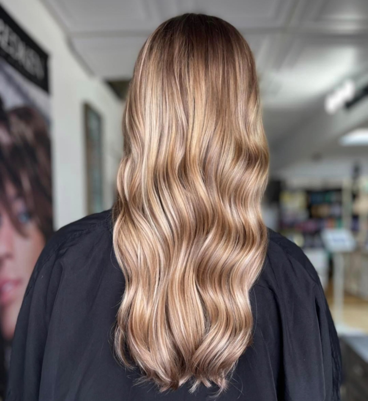 Long, wavy hair with a blend of warm blonde tones, viewed from the back. The setting suggests a salon, highlighting hairstyling expertise.