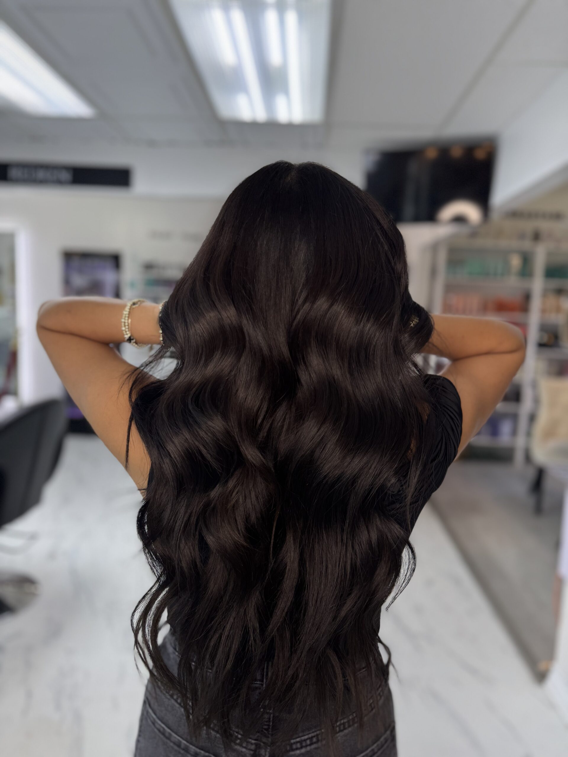 Long, wavy dark hair cascades down the back of a person in a salon setting, showcasing a stylish hairstyle perfect for inspiration.