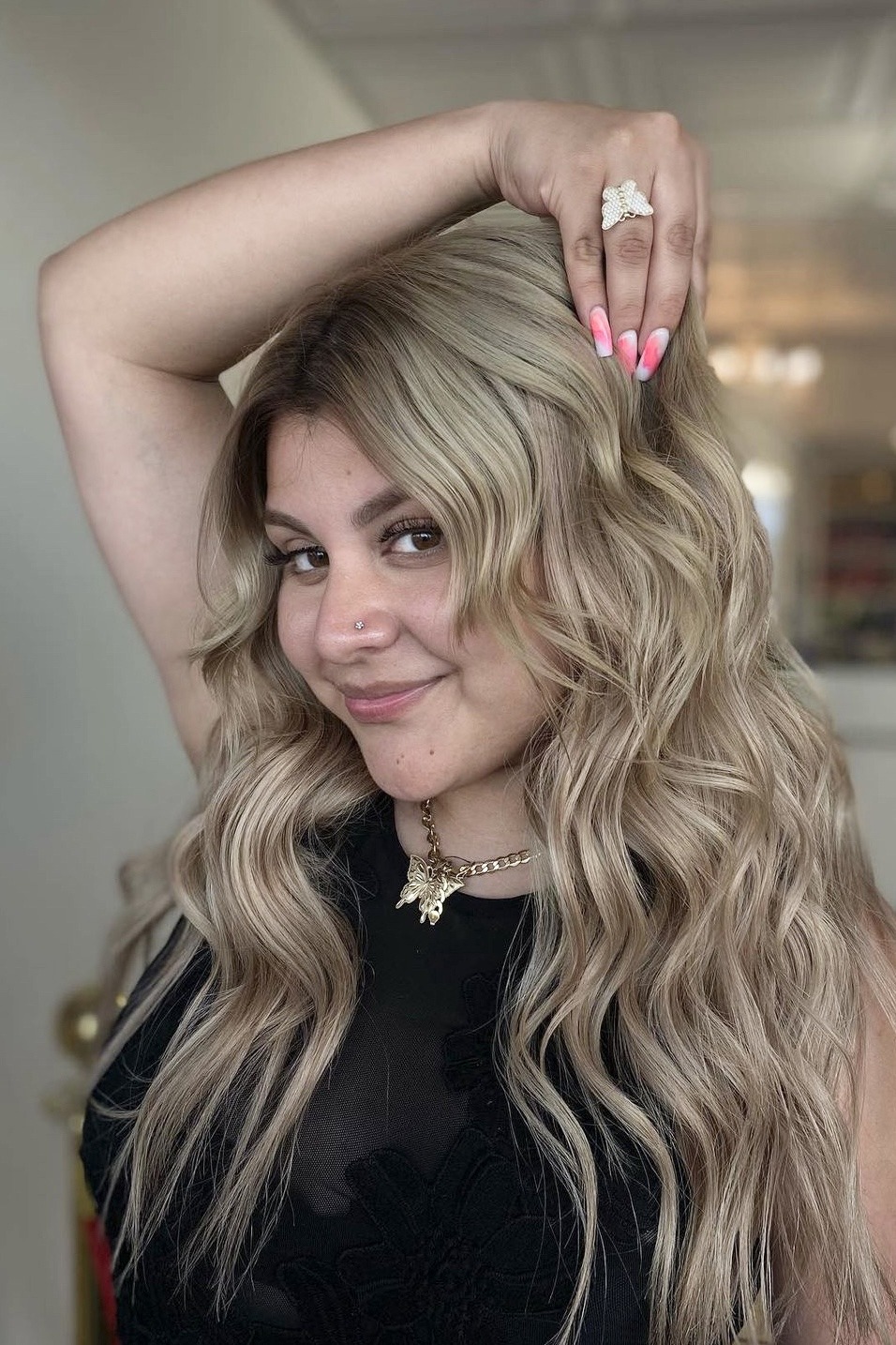 Woman with long, wavy blonde hair poses playfully, wearing a black top and statement jewelry, showcasing confidence and style in a bright setting.