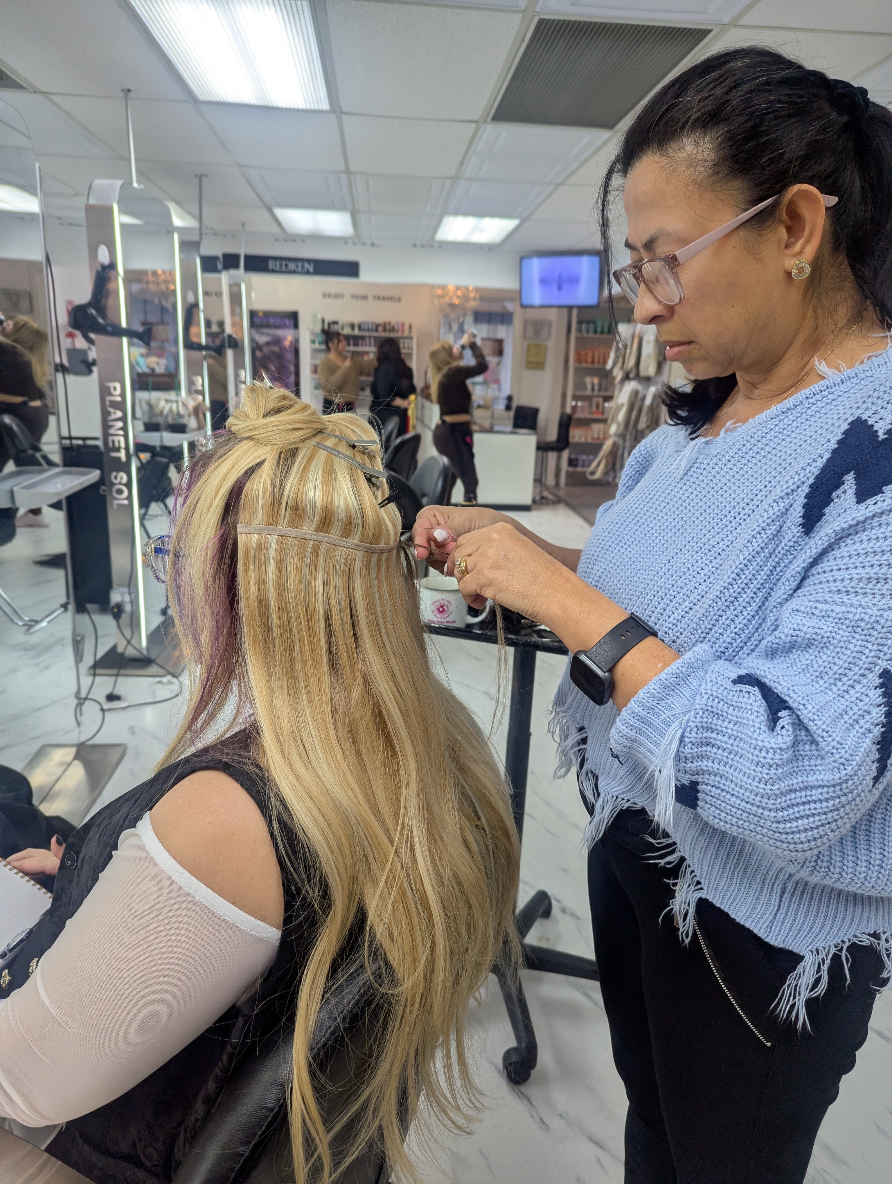 A hairstylist attaches hair extensions to a seated client's long blonde hair in a salon.