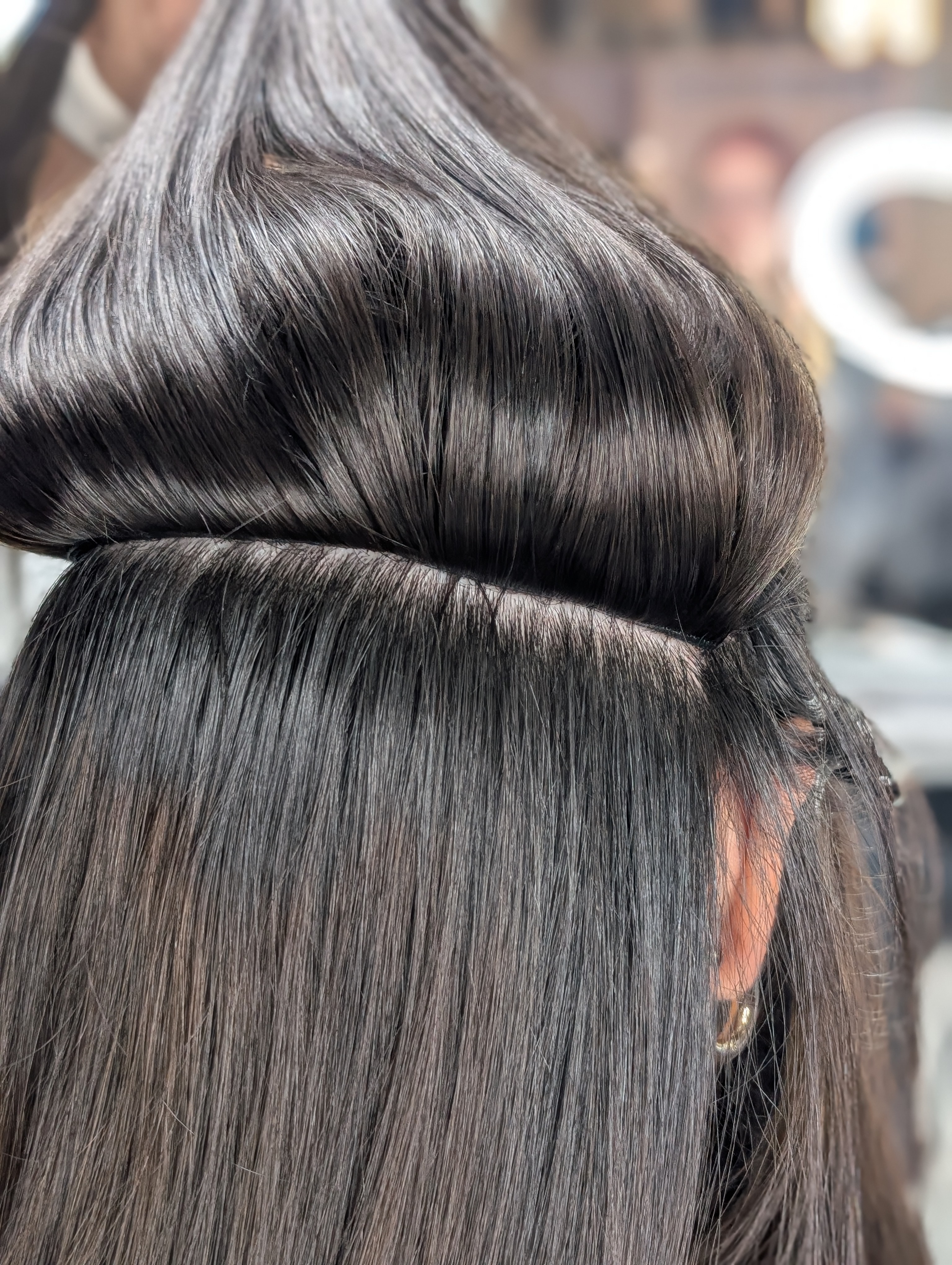 A close-up of a person's straight, dark hair sectioned and lifted at a salon, revealing the scalp and hair roots.