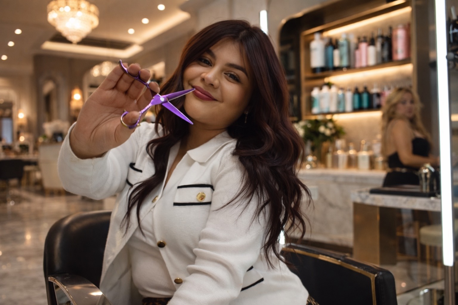 A hairstylist smiles while holding purple scissors, showcasing her work in a chic salon with elegant decor and beauty products in the background.