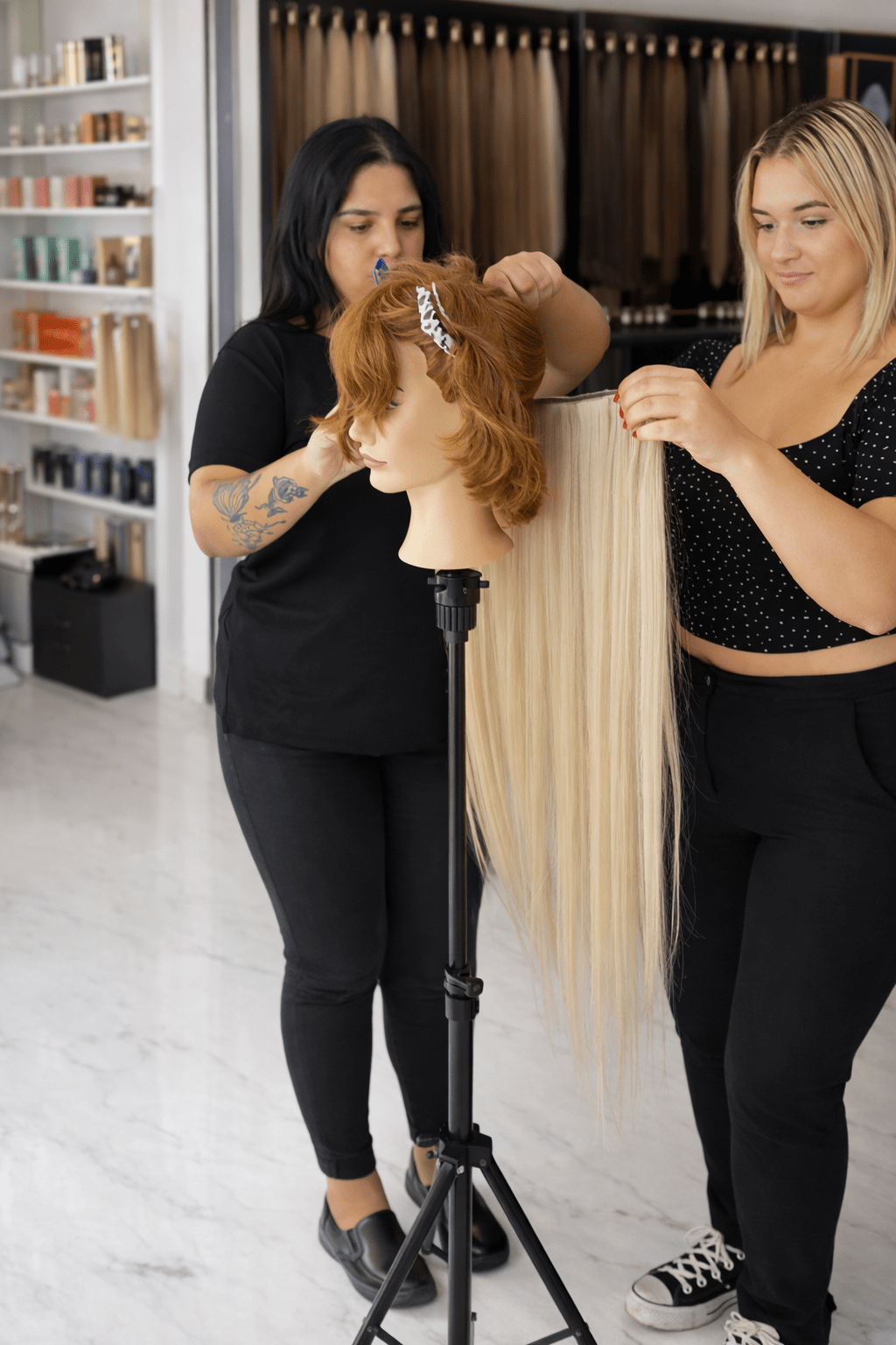 Two women work on a mannequin head with medium-length hair, adding long, blonde extensions in a hair salon setting.