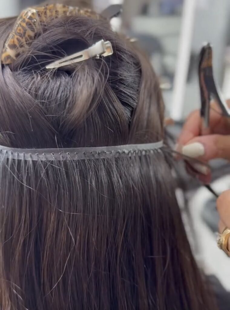 Hair extensions are being applied as a stylist uses scissors to trim. The hair is neatly sectioned, showcasing the installation process.