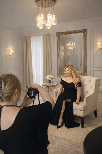 A woman in an elegant black gown poses for a photoshoot in a stylish room, while a photographer captures the moment.