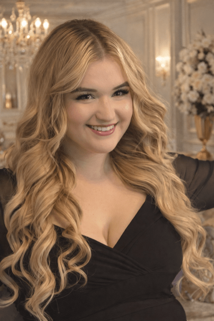 Woman with long, wavy blonde hair smiles warmly in an elegant setting, dressed in a black attire, evoking a sense of celebration and joy.